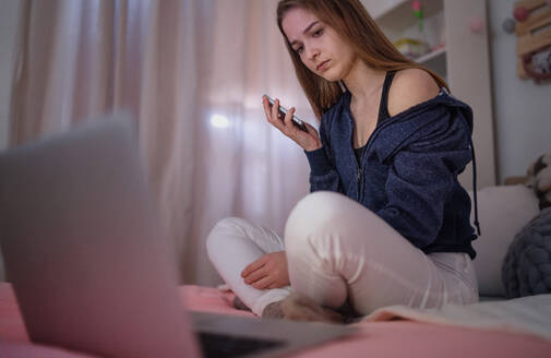 Worried young girl with smartphone sitting indoors on bed, online dating and internet abuse concept. - HPIF15070