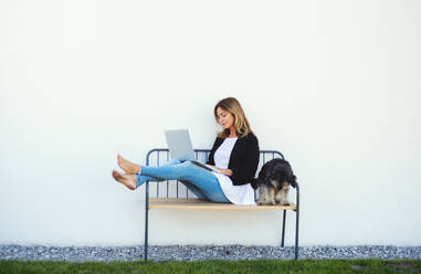 Glückliche reife Frau mit Hund bei der Arbeit im Home Office im Freien auf einer Bank, mit Laptop. - HPIF15152