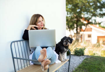 Glückliche reife Frau mit Hund bei der Arbeit im Home Office im Freien auf einer Bank, mit Laptop. - HPIF15153