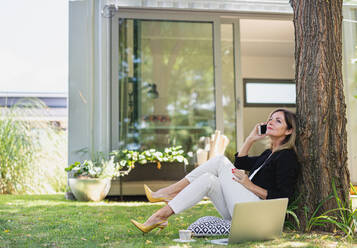 Eine reife Frau arbeitet im Heimbüro im Garten, mit Laptop und Smartphone. - HPIF15170