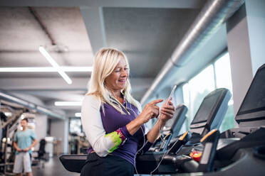 A happy senior woman with earphones and smartphone in gym doing cardio work out exercise. - HPIF15214