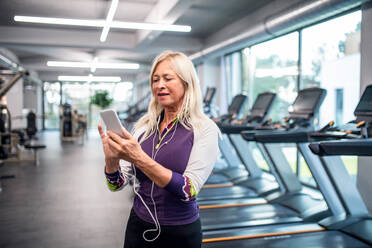 Vorderansicht einer älteren Frau mit Kopfhörern und Smartphone im Fitnessstudio, die sich nach dem Training ausruht. - HPIF15215