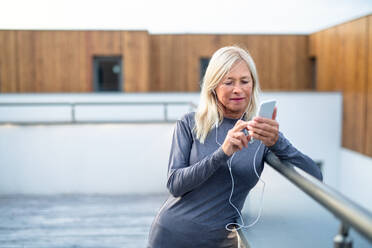 Eine ältere Frau mit Kopfhörern und Smartphone im Freien, die sich nach dem Sport ausruht. - HPIF15225