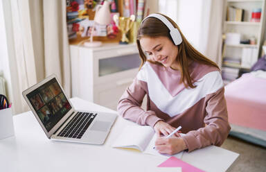 Junge glückliche College-Studentin, die zu Hause am Tisch sitzt und beim Lernen Laptop und Kopfhörer benutzt. - HPIF15289