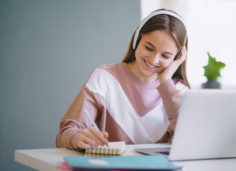 Junge glückliche College-Studentin, die zu Hause am Tisch sitzt und beim Lernen Laptop und Kopfhörer benutzt. - HPIF15304