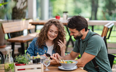 Glückliches junges Paar sitzt im Freien auf einer Restaurantterrasse und benutzt ein Smartphone. - HPIF15343