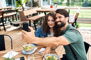 Happy couple sitting outdoors on terrace restaurant, taking selfie with smartphone. - HPIF15345