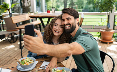 Happy couple sitting outdoors on terrace restaurant, taking selfie with smartphone. - HPIF15346