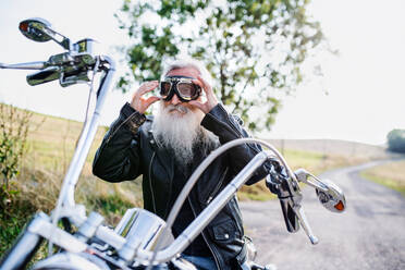 Ein älterer Mann mit Motorrad und Schutzbrille auf der Straße auf dem Lande, Raum kopieren. - HPIF15387