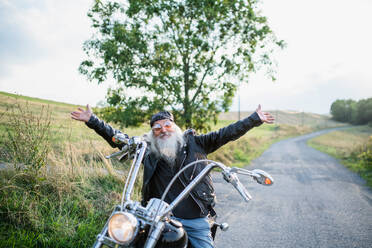 Ein älterer Mann mit Motorrad und Sonnenbrille auf dem Lande, der die Arme ausstreckt. - HPIF15404