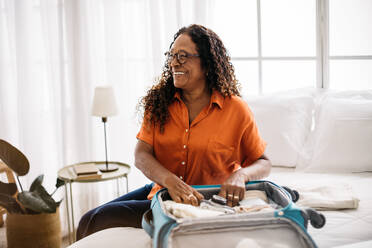 Eine Frau im Ruhestand und Weltenbummlerin sitzt auf einem Bett und lächelt, während sie ihren Koffer mit Reiseutensilien packt. Eine Frau, die sich darauf vorbereitet, neue Ziele zu erkunden und auf ihrer Reise glückliche Erinnerungen zu schaffen. - JLPSF30351