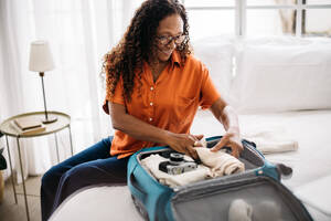 Eine Frau im Ruhestand organisiert ihr Gepäck für einen Aufenthalt in einem Luxushotel. Eine reife Frau sitzt auf einem Bett, umgeben von ihren Reiseutensilien, und lächelt, während sie ihren Koffer für eine Reise packt, die auf ihrer Wunschliste steht. - JLPSF30354