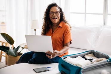 Eine Frau im Ruhestand und Jetsetterin sitzt auf dem Bett, hält ihren Laptop in der Hand und prüft online ihre Reisepläne. Eine aufgeregte reife Frau trifft Vorbereitungen, um eine Reise zu unternehmen und neue Ziele zu entdecken. - JLPSF30361