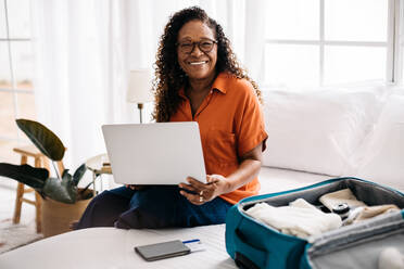 Glückliche Frau, die einen Laptop benutzt, um Online-Buchungen für eine Solo-Reise und ein Abenteuer vorzunehmen. Rentnerin, die in die Kamera lächelt, während sie auf einem Bett neben einem Koffer mit ihren Reiseutensilien sitzt. - JLPSF30362