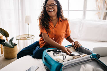 Glückliche Frau, die ihr Handgepäck für ihre Traumreise packt und ihre Reiseutensilien und -dokumente ordnet. Ältere Frau, die auf dem Bett sitzt und lächelt und sich auf eine aufregende Weltreise vorbereitet. - JLPSF30370