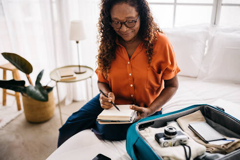 Mature woman planning her travel at home, ticking off a detailed packing list as she packs travel essentials into her suitcase. Senior woman organizing everything she needs for a successful trip. - JLPSF30371