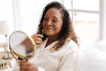 Ältere schwarze Frau, die ihre Haut mit einem Anti-Aging-Roller aus Jade pflegt. Sie schaut in den Spiegel, während sie mit dem kühlen Stein über ihr Gesicht rollt, um die Durchblutung zu fördern und Schwellungen zu reduzieren. - JLPSF30379