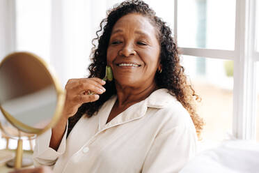 Eine alternde Frau mit melaninfarbener Haut verwendet einen Jadestein für eine Gesichtsmassage zu Hause. Mit sanftem Druck und kreisenden Bewegungen fördert sie die Lymphdrainage und baut Spannungen im Gesicht ab. - JLPSF30381