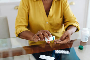 Woman sits at her table taking medication to manage her chronic disease. Senior woman following a strict treatment regimen to maintain her health, taking measures to keep her condition under control. - JLPSF30392