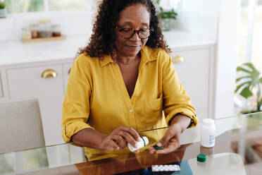 Eine chronisch kranke schwarze Frau sitzt an ihrem Küchentisch und nimmt ihre Medikamente ein. Eine ältere Frau hält sich an die ihr verordnete Behandlung, um ihre persönliche Gesundheit effizient zu verwalten. - JLPSF30393