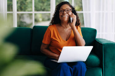Glückliche und motivierte reife Frau, die in ihrem Heimbüro sitzt und mit ihren Kunden telefoniert, während sie aus der Ferne arbeitet. Schwarze Frau, die bequem von ihrer Couch aus Geschäfte macht. - JLPSF30422