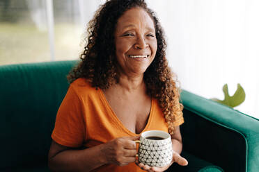 Cheerful senior woman savors her morning coffee on her couch, surrounded by the comforts of a warm home, and feeling grateful for the simple joys of life during her well-deserved retirement. - JLPSF30435