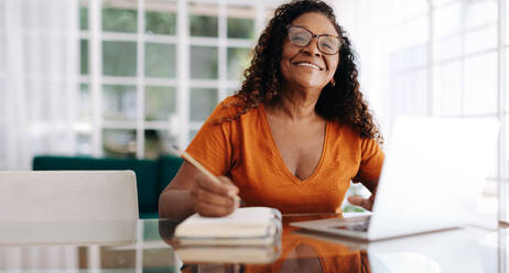 Eine zufriedene schwarze Frau, die ihren Ruhestand bequem von zu Hause aus plant. Sie sitzt mit ihrem Laptop an einem Tisch und schreibt lächelnd in ihr Tagebuch, während sie über ihre Ziele und Träume für die Zukunft nachdenkt. - JLPSF30436