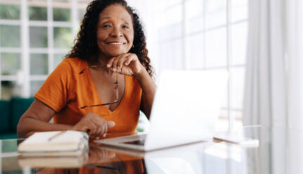 Eine erfahrene Geschäftsfrau, die in ihrem Heimbüro arbeitet, mit ihrem Laptop und einem Arbeitsjournal auf dem Tisch. Eine reife Frau, die konzentriert und produktiv bleibt, sogar in der Bequemlichkeit ihres eigenen Heims. - JLPSF30439
