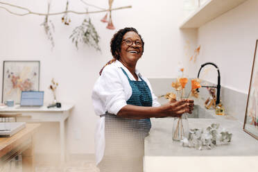 Eine ältere Unternehmerin steht mit einem fröhlichen Lächeln da und hält stolz einen handgefertigten Strauß getrockneter Blumen in ihrem kreativen Blumenarrangement-Geschäft. - JLPSF30455