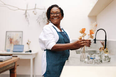 Eine Frau steht in ihrem Blumenladen und arrangiert lächelnd einen schwarzen Strauß aus Trockenblumen. Sie ist eine Unternehmerin und kreative Geschäftsinhaberin, die hart daran arbeitet, mit ihrem handgefertigten Blumenhandwerk Freude zu bereiten. - JLPSF30456