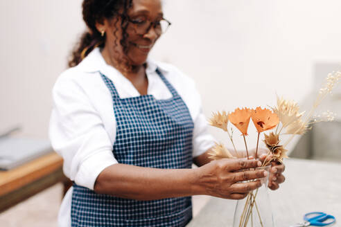 Eine glückliche Frau Ende 50, die mit handwerklichem Geschick und kreativem Gespür einen fröhlichen Strauß Trockenblumen arrangiert; eine unabhängige Unternehmerin, die ihr eigenes Blumengeschäft betreibt. - JLPSF30459