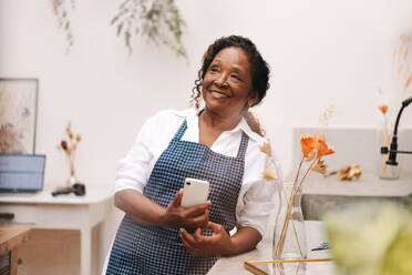 Eine ältere, selbständige Unternehmerin lächelt, während sie ihr Smartphone in einem kleinen Blumenladen benutzt. Sie arbeitet an ihrer kreativen und erfolgreichen Unternehmensgründung. - JLPSF30460