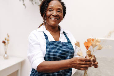 Eine ältere schwarze Frau arbeitet glücklich und professionell als Unternehmerin und kreiert in ihrem eigenen kleinen Blumenladen einen schönen Strauß Trockenblumen. - JLPSF30462