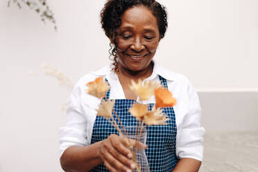 Eine schwarze Frau mit einem warmen Lächeln steht stolz in ihrem kleinen Blumenladen und arrangiert einen wunderschönen Strauß aus Trockenblumen. Sie ist Unternehmerin und kreative Floristin und fertigt handgefertigte Blumenarrangements. - JLPSF30464