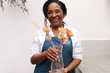 Eine erfahrene Unternehmerin steht lächelnd in ihrem kleinen Blumenladen und arrangiert mit kreativem und handwerklichem Geschick einen schönen schwarzen Strauß Trockenblumen. - JLPSF30465