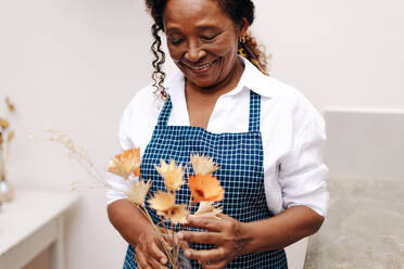 Eine ältere Geschäftsfrau steht in ihrem kleinen Blumenladen und arrangiert lächelnd einen handgefertigten Strauß aus Trockenblumen. Sie ist Unternehmerin und stolze Besitzerin dieses kreativen Geschäfts. - JLPSF30466