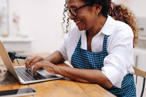 Glückliche ältere Geschäftsfrau, die einen Laptop benutzt, um die Bestellungen ihrer Kunden zu verwalten, was ihr erlaubt, organisiert und effizient zu arbeiten. - JLPSF30479