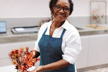 Eine Frau arrangiert einen Strauß Trockenblumen für ihr kleines Geschäft, wobei ihre Kreativität und ihr Können voll zur Geltung kommen. Eine Kunsthandwerkerin mit einer Leidenschaft für Blumendesign lächelt, während sie in ihrem Geschäft arbeitet. - JLPSF30480