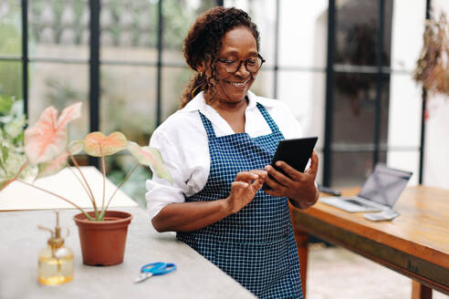 Eine glückliche Blumenhändlerin nutzt ein Tablet, um ihr kleines Unternehmen zu verwalten. Sie kann Bestellungen, Lagerbestände und Kundeninformationen an einem Ort verwalten und sich so auf die Kreation wunderschöner Blumenarrangements konzentrieren. - JLPSF30490