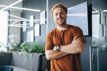 Portrait of a young business man in an office, dressed in casual clothing and smiling at the camera with confidence. Young male executive standing outside a conference room with crossed arms. - JLPPF01836