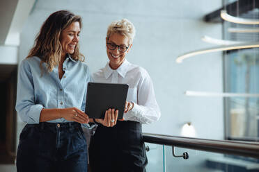 Female executive assistant holding a tablet as she shows her boss a business report indicating the company's growth and success. Happy young business woman working with a mature female professional. - JLPPF01844