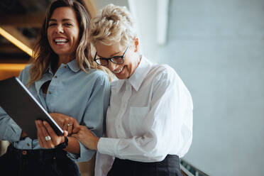 Zwei Geschäftsfrauen feiern lachend den Erfolg ihres Unternehmens, nachdem sie einen positiven Geschäftsbericht auf einem digitalen Tablet gelesen haben. Weibliche Führungskräfte arbeiten zusammen in einem Büro. - JLPPF01845