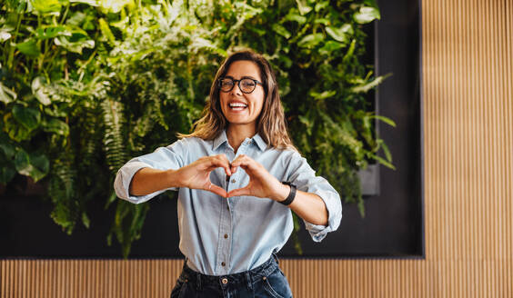 Business woman doing a heart gesture, expressing love and support for eco friendly business values. Happy female entrepreneur smiling at the camera as she stands in a sustainable work environment. - JLPPF01859