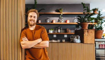 Male small business owner smiling at the camera while standing in front of his coffee shop. Young business man showing his entrepreneurship abilities as he operates a cafe. - JLPPF01860