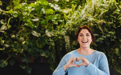 Business woman smiling and displaying a heart gesture while standing in an environmentally friendly business space. Young female executive proudly working in a green office. - JLPPF01865