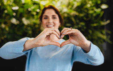 Young business woman showing her support for a sustainable and eco friendly office with a heart gesture. Professional business woman smiling and standing in a modern green office. - JLPPF01867
