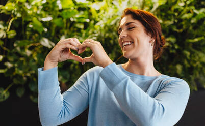 Professional business woman displaying a heart gesture and showing her support of sustainability in the workplace. Young business woman smiling as she stands in an eco friendly office. - JLPPF01868