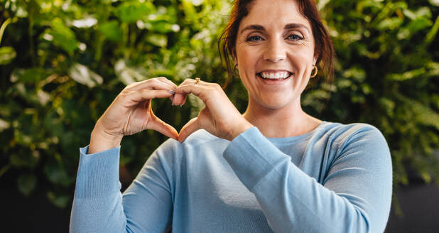 Woman showing a heart gesture and looking at the camera as she stands in a green office. Happy young business woman showing her support for sustainability in the workplace. - JLPPF01869