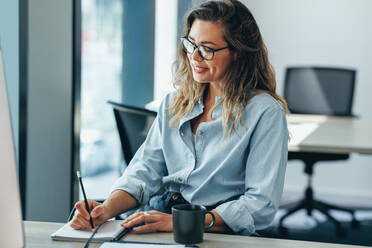 Happy business woman planning her day in the office, writing her to do list in her notebook. Young woman smiling as she works from her desk in a modern business environment. - JLPPF01877