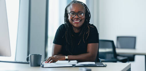 Successful business woman with dreadlocks smiles at the camera while sitting at her desk in an office. Confident and experienced African business woman working in a startup - JLPPF01887